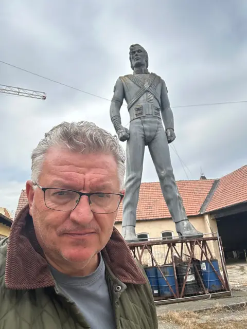 Where are Michael Jackson's giant HIStory statues 30 years later? Franz Josef Zika A man with glasses and silver hair is taking a selfie in front of a Michael Jackson statue perched on a base made of oil drums. Parts of the statue look brown and stained white.
