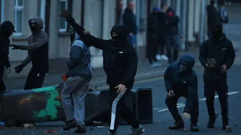 Police deploy more officers after 'mob violence' Getty Images Six men wear black coats and black tracksuit bottoms. They all wear black balaclavas as they throw stones, bricks and other objects. A group of people, out of focus, can be viewed looking on in the background. Two black wheelie bins lie beside the individuals throwing the rocks.