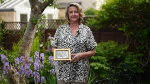 More pets being put down due to rising costs, BBC told Caroline is in her garden. She is holding a picture of Ozzie, a cat, in a golden frame.