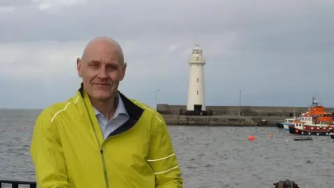 Second arrest in Donaghadee murder inquiry Mark Brooks in yellow jacket standing in front of the harbour and lighthouse in Donaghadee, with an orange, white and blue boat in the far right corner.