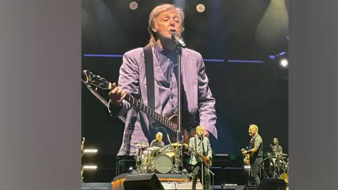 Bruce Springsteen and Paul McCartney in surprise Liverpool duet bruce2023tour /X Sir Paul McCartney wearing a grey blazer, stands in front of a microphone, holding a guitar, on a huge stage. Bruce Springsteen is to his left, smiling at him, as he holds a guitar. A man is on the drums behind them.