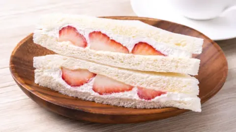 Japanese fruit sando hits high streets Promo_Link/Getty Images A strawberry and cream sandwich in white bread, served on a wooden plate on top of a table