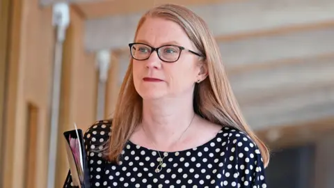 Scotland to recover winter fuel payment from better-off OAPs Getty Images Shirley-Anne Somerville - a a woman with glasses, shoulder length auburn and wearing a black dress with white polka dots - walks in a corridor at Holyrood while carrying a folder