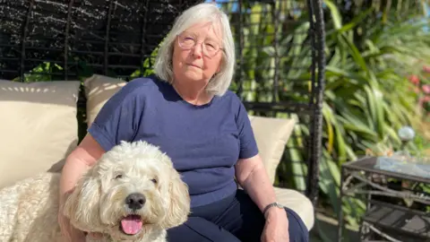 More pets being put down due to rising costs, BBC told BBC Avril is sitting outside in a blue top. She is sitting on a black wicker chair. Beside her is Dougie who is a white dog.