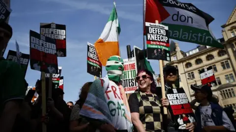 Rapper released on unconditional bail over terror charge EPA A crowd of people holding placards saying 'Defend Kneecap', and holding Palestinian flags and Irish flags outside the court in London