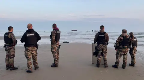 Smugglers deploy 'taxi boats' to collect migrants off the French coast Marianne Baisnée/BBC Six French gendarmes stand facing the sea, their backs to the camera. In the distance you can see a large inflatable boat close to where dozens of people are gathered in waist-deep water
