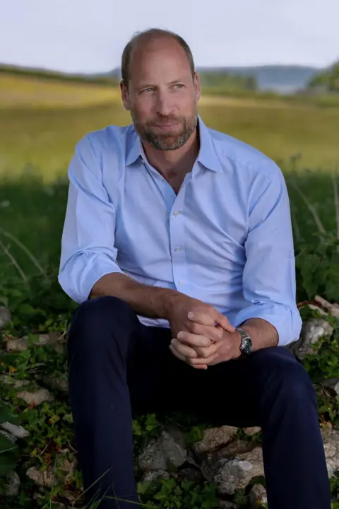 Prince William celebrates 43rd birthday with puppy photo Kensington Palace Prince of Wales, wearing a light blue shirt, looks into the distance while sitting on a stone wall with a meadow behind him