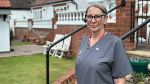 Covid inquiry begins examination of impact on social care sector Maureen Lewis, wearing a grey tunic and red lanyard, standing in the garden of St Ives Lodge care home. She has her hair tied back and is wearing glasses.
