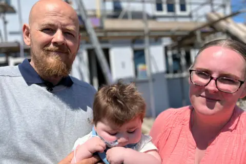 Parents' worldwide search for babies with son's rare condition Family photo Amanda and Nick are holding Jack in front of their house which is covered in scaffolding. Amanda has a pink long-sleeved top, wears glasses and has her fair hair tied back in a pony tail. Nick has a beard and is wearing a grey jumper and jeans. Jack is in a blue baby grow and has a feeding tube going up his nose.