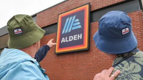 Aldi in Oasis rebrand as homage to band ahead of Manchester gigs PA Media Two men wearing bucket hats, one green and one blue, stand with their back to the camera and point at the Aldi sign, which features the shop's orange and red bordered blue logo and the word Aldeh, on the side of one their brick built store