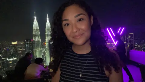 Asos customers banned over returns say it is 'slap in the face' A woman smiles in the camera wearing a black sleeveless dress. Neon purple lights are visible in the background