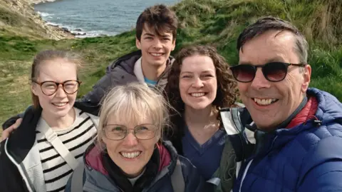World-first 'Trojan horse' therapy to be given on NHS Paul Silvester Paul and his family - wife, two daughters and son - all smiling while they are out for a hike with cliffs and the sea in the background