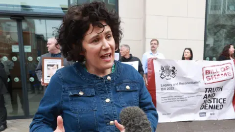 Ballymena sees second night of disorder with missiles thrown at police PA A woman with wiry black hair speaking into a mic, she has a denimn jacket on. Behind her is protestors with a white banner.