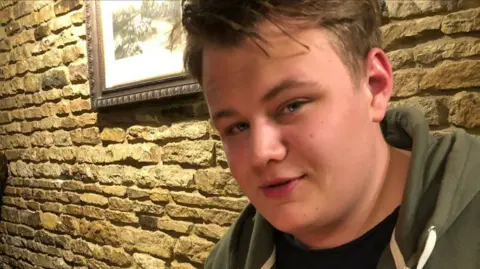Foreign Office faces inquiry over its handling of Harry Dunn case Dunn family Harry Dunn wears a green hoody. He sits in front of a stone wall with a framed picture on it.