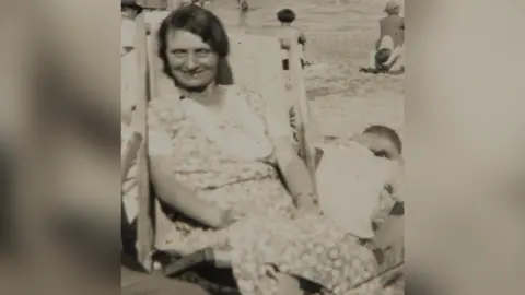 Handout Old photograph of Louisa Dunne. She has dark hair and is sat in a deck chair wearing a floral dress. She is looking at the camera and smiling.