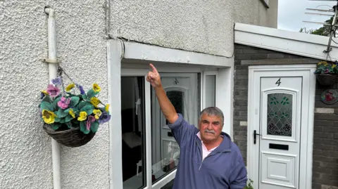 'The Heads of the Valleys road swallowed part of my garden' Daryl Wilkins pointing to a crack above his head in the external white render of his home. He is wearing a blue sweatshirt and has grey hair and a grey moustache. He is standing in front of a window, with his front door to the right and a hanging basket to his left.