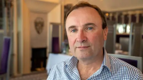 Fraudster jailed for selling fake 'Scottish-grown tea' A man with short, brown hair looks at the camera. He is wearing a blue and white checked shirt and appears to be sitting in a restaurant