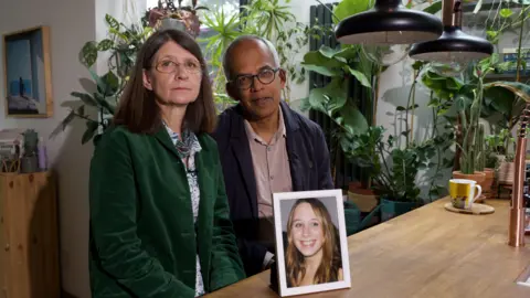 NHS trust and medic guilty of failings Jane and Max Figueiredo sitting at a table looking at the camera, with a framed, smiling photo of Alice, a woman with long blonde in the foreground. Jane is wearing a green blazer, glasses and has shoulder-length dark hair. Max is balding, wears glasses, and wears a dark blue blazer. There are many plants in the background.