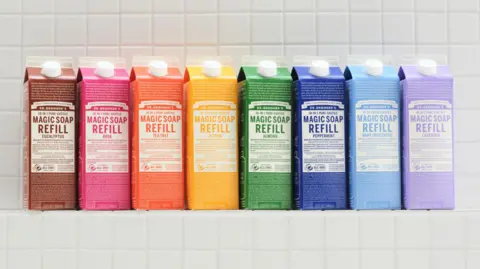 The global environmental award hit by accusations of greenwashing Dr Bronner's A row of eight refill packs of Dr Bronner's Magic Soap in many bright colours lined up on a white-tiled shelf.