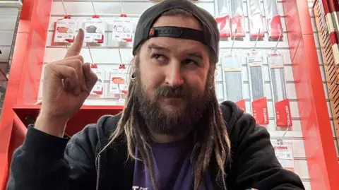 Norwich music store closure brings fear for industry's future Dan Moss Dan Moss is a man with shoulder length dreadlocks. He is looking to the left of the image. On the left of the image, his finger is pointing upwards. He is wearing a dark jacket over a purple t-shirt, and is wearing a backwards baseball cap.