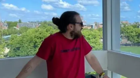 Norwich music store closure brings fear for industry's future James Wickham Dylan Barber is a man, looking to the right of the image. He is wearing sunglasses, and has a beard. His hair is tied up into a bun. He is wearing a bright red tshirt, and behind him there is a white ledge, and green trees behind it.