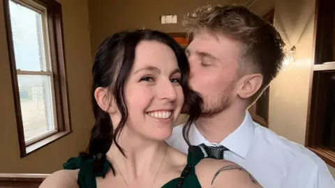 'It doesn't matter that my boyfriend is 5ft 6in"' Ashley, a young woman with long brown hair wearing a grey top, takes a selfie with Joe, a young man with blonde hair wearing a white shirt and grey tie who is kissing her on the cheek.
