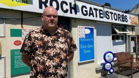 Post Office criminal trials may not start until 2028, says police chief Tim Brentnall, with glasses and shaven head, stands with his hands behind his back wearing a brown, black and white Hawaiian-style shirt. In the background is his former store and Post Office, with a sign saying "Roch Gate Stores". A post office logo is visible.