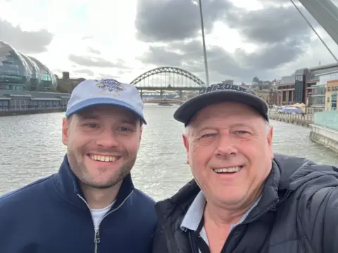 Dad gave me a kidney so I can have my dream wedding Brian Innes Kieran is smiling at the camera. He is wearing a dark blue fleece jacket and cap. His dad, Brian, is also wearing a cap and puffa jacket. The Newcastle bridge and Tyne River are in the background.