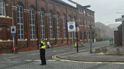 Ship on fire at Hull's Albert Dock as roads and shops are closed Amber Gash/BBC A police officer wearing black trousers and a yellow high-vis vest stands in the centre of a road. A large brick building is in the background, surrounded by hazy grey smoke.