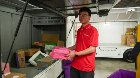 China's driverless lorries hope to expand BBC/Joyce Liu Man wearing red shirt loads parcel onto the back of a truck