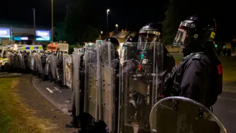 Families hid in attics during 'race motivated' disorder, says chief constable PA Media A line of police in riot gear. Their faces are covered.