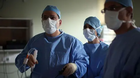 Hundreds of NHS quangos to be scrapped in 10-year health strategy Getty Images A surgeon and his theatre team perform key hole surgery to remove a gallbladder at the Queen Elizabeth hospital in Birmingham. The doctors are all wearing blue overalls and white masks and staring off camera. The main surgeon is holding a medical device.