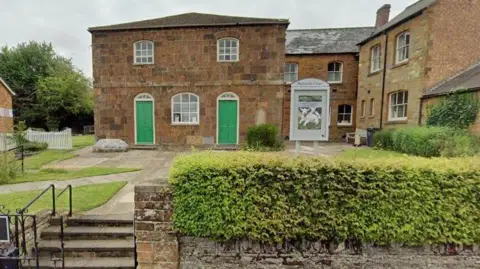 Cost of living roadshows to tour West Northamptonshire Google Two-storey stone-built chapel with two green doors on either side of a arched window on the ground floor and two arched windows above, with small square pains of glass separated by white frames. There is a grassed area in front, criss-crossed by paths and another stone building to the right. There is a hedge in the foreground and some stone steps with railings.