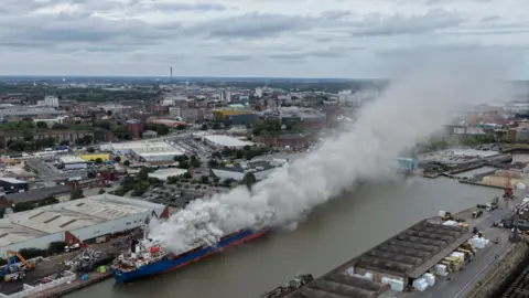 Ship on fire at Hull's Albert Dock as roads and shops are closed Dale Baxter/BBC A blue and red ship in a dock which grey smoke billowing out. The Hull skyline is in the distance.
