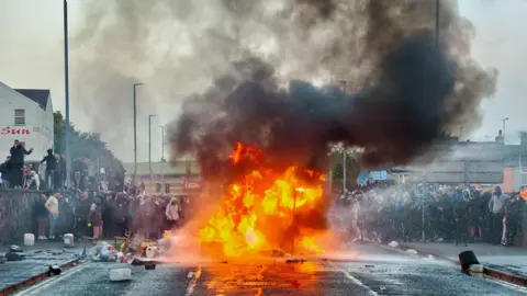Ballymena sees second night of disorder with missiles thrown at police Pacemaker Bright orange flames are in the foreground of a blocked road with a large crowd of people standing close behind.