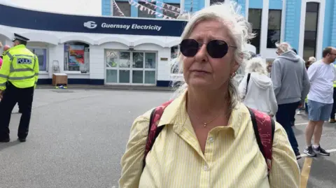 'We sit in the dark to save money on electricity in Guernsey' A woman in sunglasses and a yellow sirt in front of the Guernsey Electricity headquarters, with police and protestors behind her.