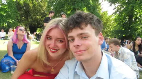 Our sister died because of our mum's cancer conspiracy theories, say brothers Ander Harris Paloma Shemirani, with a streak of pink in her hair and red lipstick, wearing a red off the shoulder outfit, leaning in close to Ander Harris, a white man with brown hair, tousled on top, wearing a light coloured collared shirt. They are both smiling as they sit on the grass, surrounded by trees and other young people, presumably students.