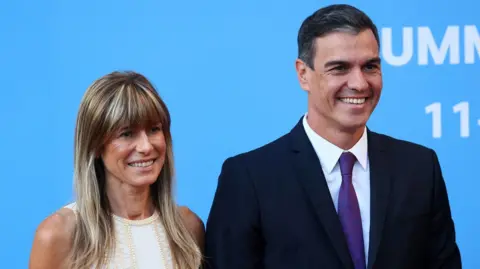 Embattled Sánchez resists clamour for resignation Reuters Sánchez and his wife Begoña Gómez are smiling with a blue background. Sánchez is wearing a suit and Gómez a white dress.