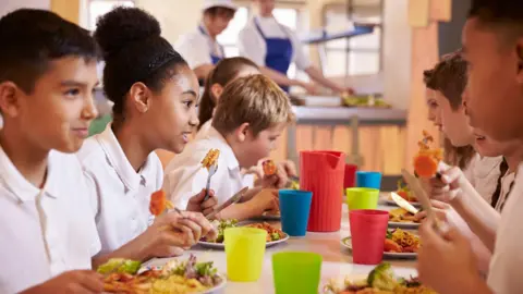 Getty Images Several primary school pupils sit at a canteen table eating a meal. They are all wearing white uniform polo shirts and are smiling at one another. Alongside the food in front of them are multi-coloured drinking cups and a jug.