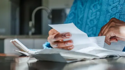 What are the Pip and universal credit changes and who will be affected? Getty Images Close up shot of a person looking at bills and receipts on a table next to a calculator in their kitchen. They are wearing a blue jumper and the kitchen sink is in the background.
