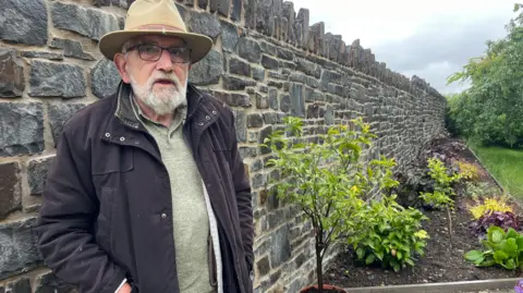 'The Heads of the Valleys road swallowed part of my garden' BBC John Watkeys standing against his new stone garden wall which is about head height. He is wearing a fawn-coloured hat and glasses with a black coat and has a white beard. A flower bed can be seen to the right of the wall and grass to the right of that.
