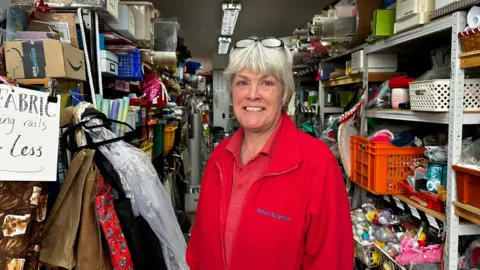 NHS and education named as local priorities A woman in a red zip up jumper that says 'Saltash Scrapstore' and a red top. She has short hair and glasses on her head. She is smiling at the camera. Around her is various items of scrap on shelves.