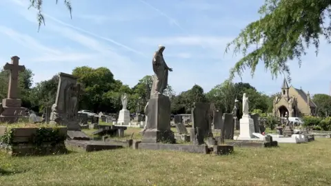 London funeral costs highest in UK, report finds A cemetery with a variety of tombstones, stone crosses, and statues, including religious figures. In the background stands a small chapel or church with a pointed roof, surrounded by trees under a clear sky.