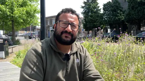 Why is Bristol's birth rate falling faster than other UK cities? Courtney Young sitting on a bench nearby a road with a patch of grass and some flowers. He is wearing a khaki green fleece and has black rimmed glasses and curly hair.