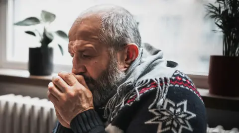Scotland to recover winter fuel payment from better-off OAPs Getty Images A man in a Christmas jumper huddles for warmth next to a heater