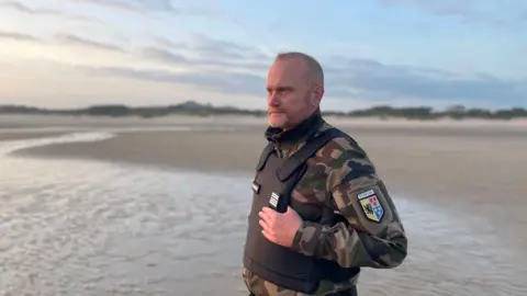 Smugglers deploy 'taxi boats' to collect migrants off the French coast Marianne Baisnée/BBC A man in his fifties dressed in police uniform, with a bullet-proof vest, stands on the sand of a beach looking out to see