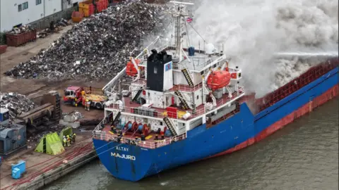 Ship on fire at Hull's Albert Dock as roads and shops are closed Dale Baxter/BBC A blue and red ship in a dock with grey smoke billowing out. The Hull skyline is in the distance.