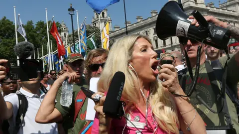 Our sister died because of our mum's cancer conspiracy theories, say brothers Getty Images Kate Shemirani, who has long blond hair and wears a pink outfit with what appears to be a stethoscope around her neck, at an anti-lockdown protest in 2021. A man nearby holds up a megaphone speaker as she speaks into the microphone part, which she holds in her hand. Behind her are flags on what looks like Parliament Square, along with crowds of people, some holding up cameras which they are pointing at the press photographer.