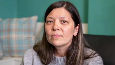 Hopes to open NHS ketamine clinic for treatment resistant depression A photo of Alex looking directly at the camera. She has long brown hair. She is wearing a grey woollen jumper. The blurred background is a settee with patterned cushions