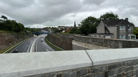 'The Heads of the Valleys road swallowed part of my garden' A view from a bridge down onto the Heads of the Valleys dual carriageway. On the right hand side is a large stone wall, and John's house on a platform to the right of the wall. It is a cloudy day.